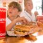 A red Five Guys soft drink cup is the focus in the foreground, as children enjoy their meals in the background. A red Five Guys soft drink cup is the focus in the foreground, as children enjoy their meals in the background.