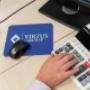 Person typing on calculator next to blue Virtus Group mousepad on desk Person typing on calculator next to blue Virtus Group mousepad on desk